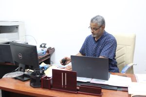 Man wearing glasses sits at a desk with a laptop, holding a headset in an office setting with a printer and papers nearby
