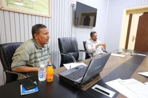 4C545AB1-D7C8-4129-BE15-AE07C3A90A09 - جامعة سبها | Sebha University Two men seated at a conference table in a meeting room, one speaking while the other listens, with a laptop and papers on the table.