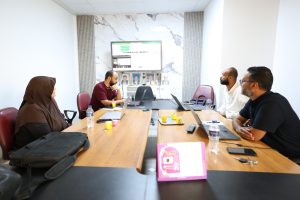 51B37536-00CF-4F08-AC95-378F56521EB1 - جامعة سبها | Sebha University Group of four professionals in a boardroom around a wooden table with laptops and water bottles, a large monitor on a marble wall behind them.