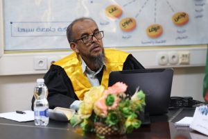 5301580C-9201-4662-A6F2-D978838FB44A - جامعة سبها | Sebha University Senior academic in yellow robe sits at a desk with a laptop, flowers, and a water bottle during a meeting or presentation.