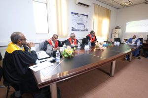 544A8D27-AB05-48CB-B99A-233C0C311CA9 - جامعة سبها | Sebha University Five academics in robes and red sashes sit at a long conference table during a formal meeting, with a projector screen and floral centerpieces nearby.