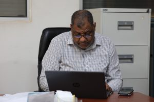 Older man in glasses works on a laptop at his desk in an office setting behind a filing cabinet.