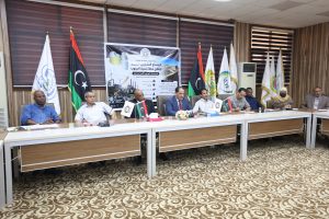 5C9DE023-CB2D-404B-B3AD-02D0C716AB30 - جامعة سبها | Sebha University Panel of officials seated at a long conference table with flags on the sides and a banner backdrop behind them.