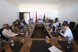 5E990143-6A5E-4BA0-A2D9-23166E51B1F0 - جامعة سبها | Sebha University Group of people seated around a long conference table in a meeting room, with flags and a banner in the background.