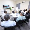 Presenter stands at the front of a bright conference room as a screen shows a slide while colleagues sit around a round table.