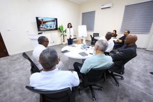 Presenter stands at the front of a bright conference room as a screen shows a slide while colleagues sit around a round table.