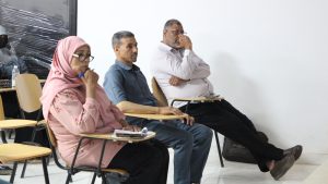 Three adults sit in a classroom or meeting room, listening attentively: a woman in a pink hijab and glasses in the foreground, flanked by two men.