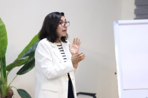 Woman in a white blazer and striped shirt explains a point, gesturing with her hands beside a whiteboard in a bright room.