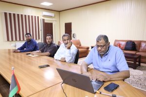 68B3CC7B-43D3-4278-91DE-FA7B9381F16D - جامعة سبها | Sebha University Four men seated along a conference table in a meeting room, laptops and a smartphone on the table, flag in foreground.