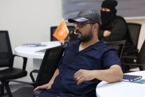 Man wearing a navy shirt, cap, and glasses seated at a round table in an office; a woman in a niqab sits in the background.