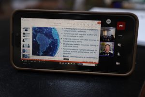 Phone screen shows a video conference with a slide deck featuring a globe image and bullet points; two speakers appear in video thumbnails on the right.
