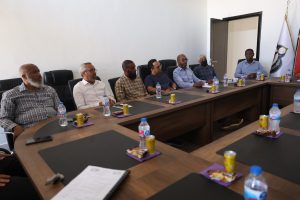 6CE6F652-89B5-497F-BF2A-C97A0174DE58 - جامعة سبها | Sebha University Group of men seated along a long conference table during a meeting, with water bottles and snacks on purple napkins nearby.