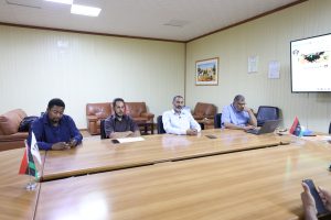 6ED2D16D-1C72-4125-8F45-C5E43E13EB2F - جامعة سبها | Sebha University Four men sit around a long wooden conference table in a beige-walled meeting room, with small flags and a large wall screen visible to the right.