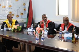 6F032627-4468-4847-9D71-1482FEAC9EF2 - جامعة سبها | Sebha University Three men in academic robes sit at a conference table with water bottles and flowers, Palestine flag behind them, during a formal meeting.