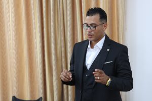70FF5FA6-1EC6-43D1-964C-A3FFE7DDCFBE - جامعة سبها | Sebha University Man in a dark suit and white shirt, wearing glasses, gesturing as he speaks in front of gold-patterned curtains.