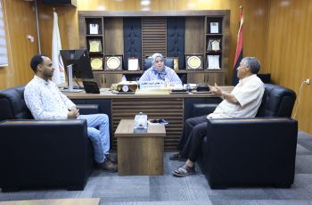 Three people sit in a formal office around a wooden desk; a woman in a headscarf sits center with two men nearby and awards on shelves behind her.