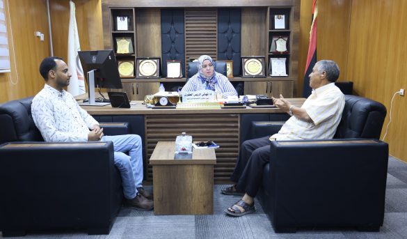 Three people sit in a formal office around a wooden desk; a woman in a headscarf sits center with two men nearby and awards on shelves behind her.