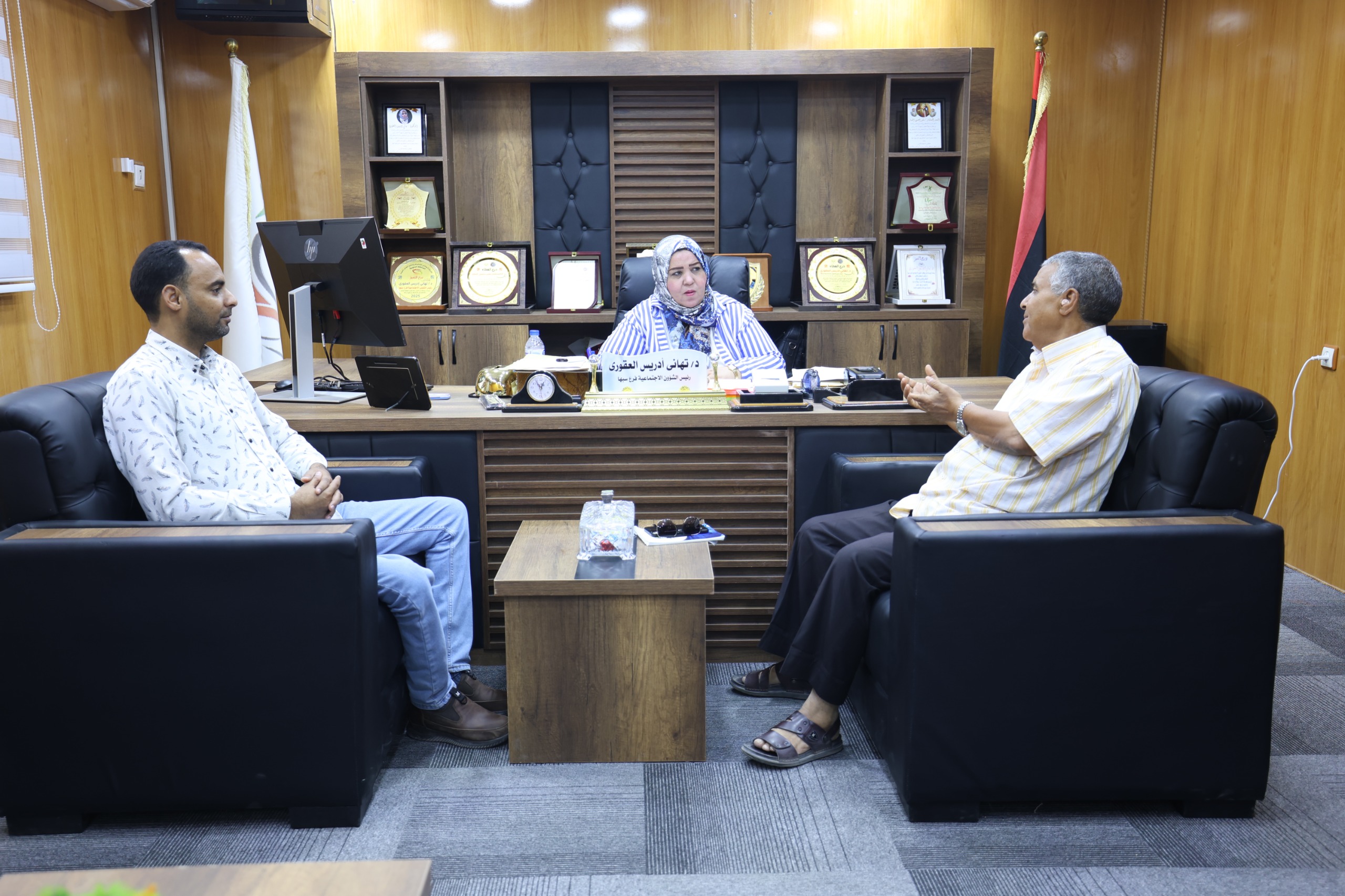 Three people sit in a formal office around a wooden desk; a woman in a headscarf sits center with two men nearby and awards on shelves behind her.