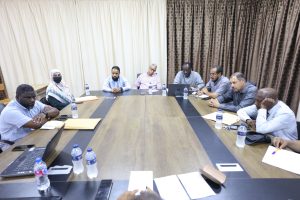 Group of eight people seated around a long conference table with laptops, papers, and water bottles during a meeting.
