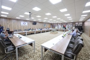 736D4E9C-3A65-4CFB-ADD1-F671C4DFC003 - جامعة سبها | Sebha University Conference room with attendees seated around a U-shaped table, flags lined up at the back, and a Sebha University banner on a wood-paneled wall