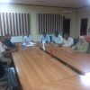 Eight people sit around a long wooden conference table in a meeting room, with small flags and papers in the center.