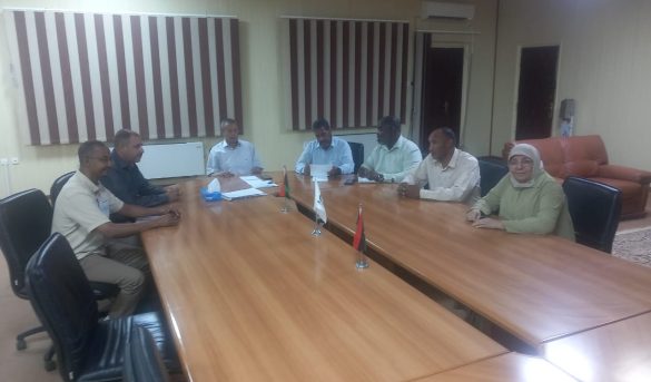 7404A950-D040-4C46-9CB6-4FB0D4BB2ECB - جامعة سبها | Sebha University Eight people sit around a long wooden conference table in a meeting room, with small flags and papers in the center.
