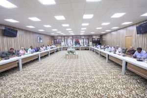 Large conference room with a U-shaped table and many attendees seated along the sides; flags line the back wall and a central flower arrangement sits on a pedestal.
