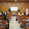 Speaker presenting to a classroom audience in a wood-paneled lecture hall with a projector screen behind him, many attendees wearing hijabs.