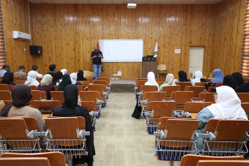 7962E8DC-626A-4CA0-9850-D17165FE4041 - جامعة سبها | Sebha University Speaker presenting to a classroom audience in a wood-paneled lecture hall with a projector screen behind him, many attendees wearing hijabs.