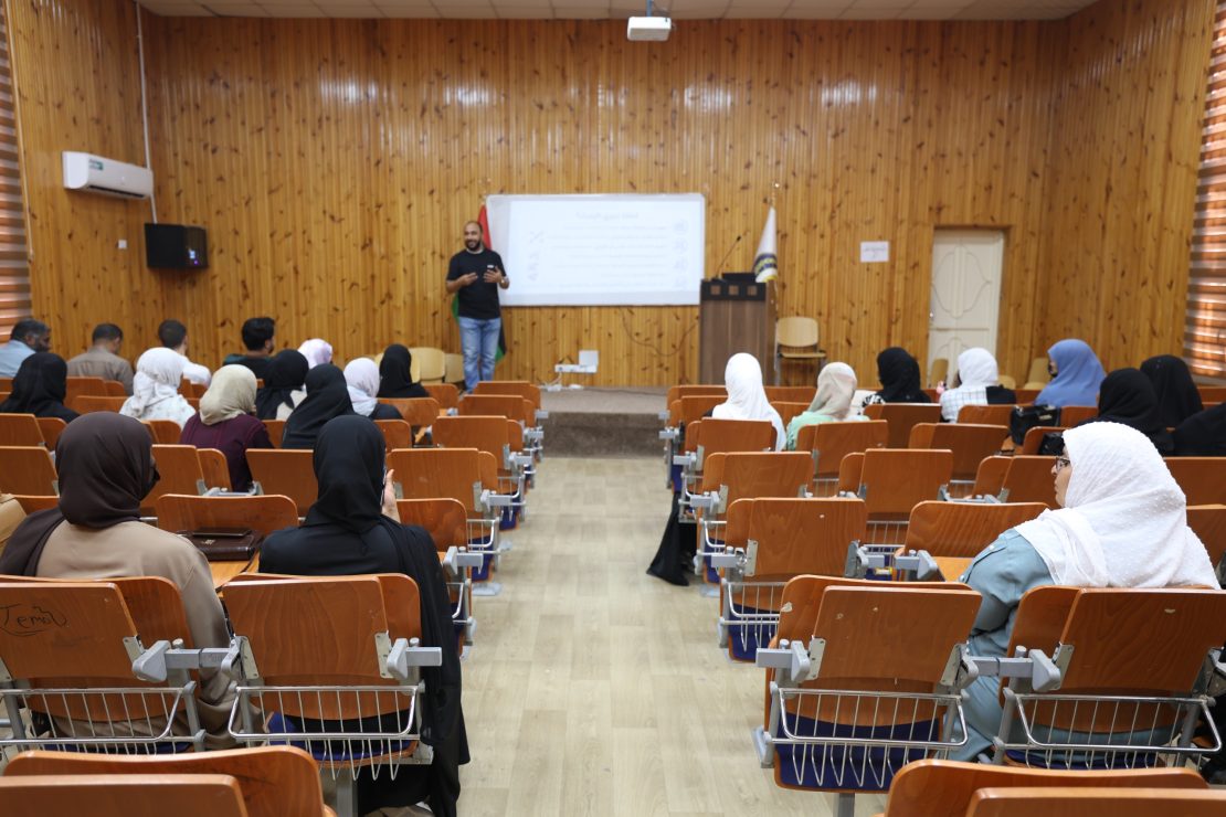 Speaker presenting to a classroom audience in a wood-paneled lecture hall with a projector screen behind him, many attendees wearing hijabs.