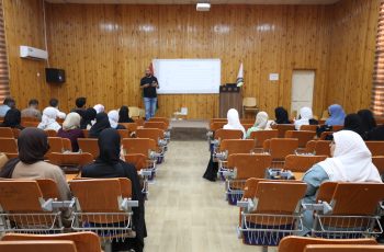 Speaker presenting to a classroom audience in a wood-paneled lecture hall with a projector screen behind him, many attendees wearing hijabs.