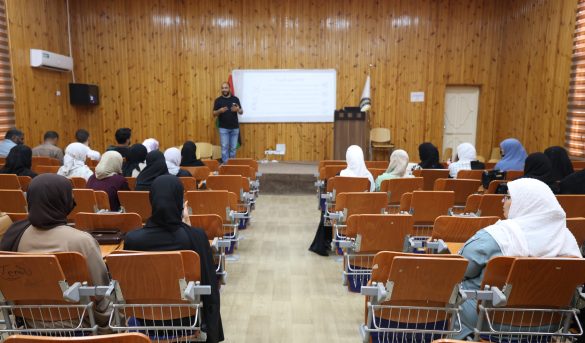 Speaker presenting to a classroom audience in a wood-paneled lecture hall with a projector screen behind him, many attendees wearing hijabs.
