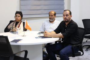 Three adults sit around a round white table in a meeting room, looking attentive as they discuss (orange flag and notebooks on the table).