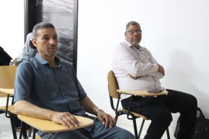 Two men seated in a bright room on wooden desks with attached tablet-style surfaces, listening in a panel-style setting – one in a blue shirt foreground, the other in a striped shirt and glasses to the right.