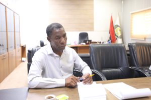 7DB8750D-A109-4FB4-A6AE-AA5B07E2C381 - جامعة سبها | Sebha University Man in a white shirt sits at a conference table, signing papers with pens in an office setting with cabinets and flags in the background.
