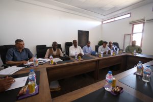 Group of men in a meeting room seated along a long conference table with papers, notebooks, and water bottles in front of them.