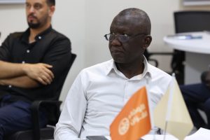 Man in a white shirt and glasses sits at a conference table with orange and pale flags in the foreground; another man in black sits in the background in a meeting room.