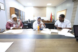 7FEE40AD-52D3-488E-8E0F-5C00C83FEF1B - جامعة سبها | Sebha University Three colleagues sit at a conference table in a boardroom, two women wearing hijabs and a man in a white shirt, discussing documents.