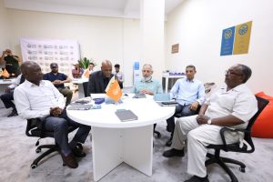 A group of men sit around a white round table in a bright meeting room, with orange flags and notebooks on the table, discussing.