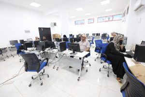 8398A4D5-E8B7-480D-914C-D103BDE3582B - جامعة سبها | Sebha University Computer lab with desks, monitors, and blue chairs as people work at desktop PCs in a bright, white room.
