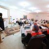 Woman in a hijab presenting at the front of a conference room with a projector screen behind her.