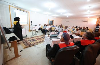 Woman in a hijab presenting at the front of a conference room with a projector screen behind her.