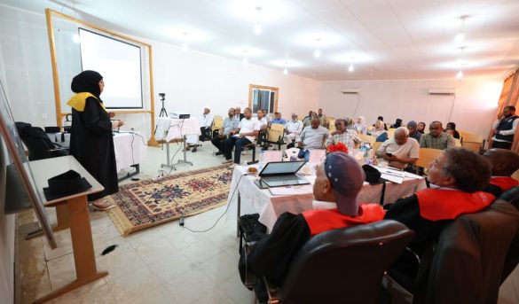 Woman in a hijab presenting at the front of a conference room with a projector screen behind her.