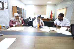 860D535F-F1D4-447B-A746-28729DB78516 - جامعة سبها | Sebha University Three professionals sit around a conference table with papers and drinks, in a meeting room.