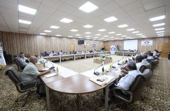 Large conference room with a U-shaped table setup and 20+ attendees discussing, flags and a SEIBA University banner in the background.