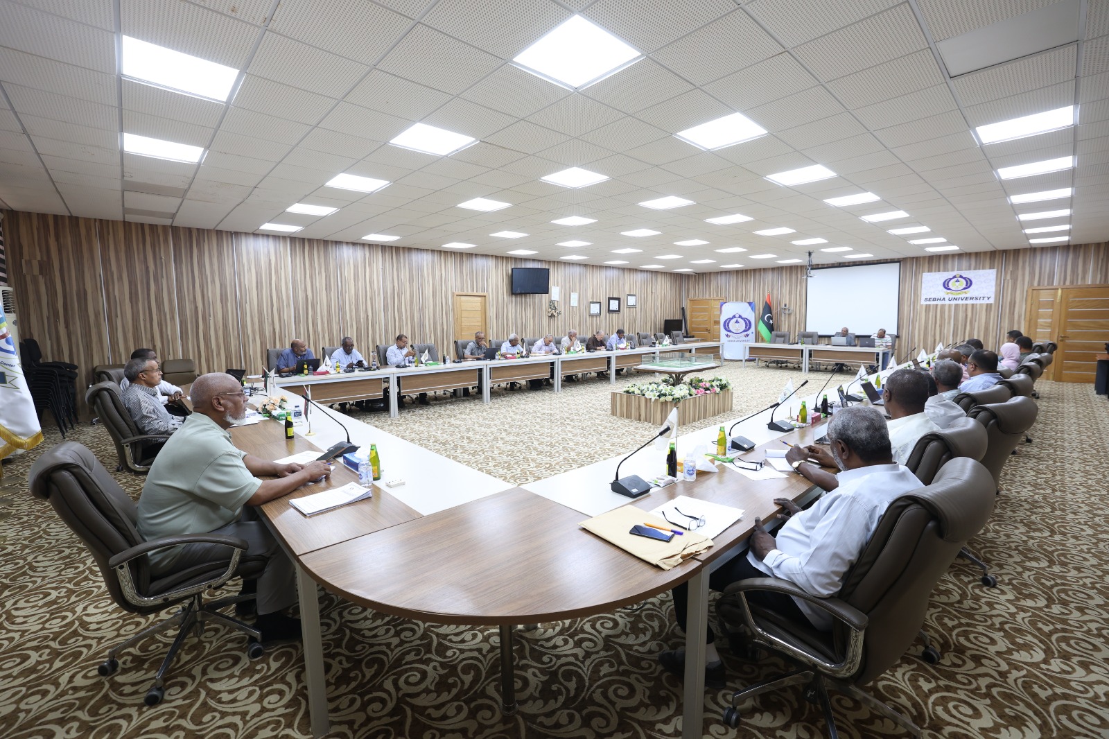 Large conference room with a U-shaped table setup and 20+ attendees discussing, flags and a SEIBA University banner in the background.