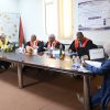 Formal panel meeting around a long table with five people in robes, banner on the wall, and flag in the corner.