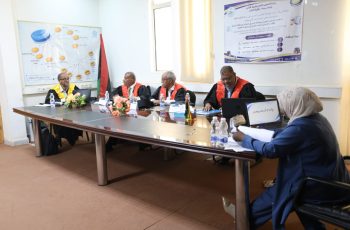 Formal panel meeting around a long table with five people in robes, banner on the wall, and flag in the corner.