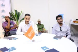 Four men sit around a white round table in a meeting room, with notebooks and two flags on the table and a plant in the background.
