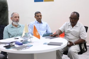 Three men sit around a round table in a meeting room, reviewing documents with small flags on the table.